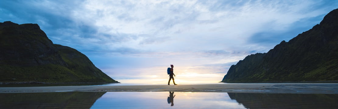 A lone person walking across a reflective shoreline between mountains at sunrise, symbolizing the six must-know principles that help new investors begin their journey with confidence and a strong foundation
